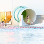 Ein Mädchen steigt von einer Wasserrutsche in ein Schwimmbecken, während drei Personen in der Nähe schwimmen; leere gelbe Stühle säumen den Beckenrand im Hintergrund.