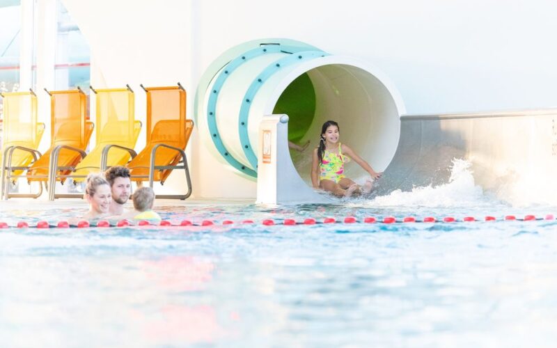 Ein Mädchen steigt von einer Wasserrutsche in ein Schwimmbecken, während drei Personen in der Nähe schwimmen; leere gelbe Stühle säumen den Beckenrand im Hintergrund.