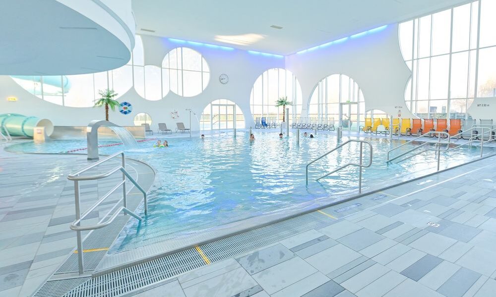 Hallenbad mit mehreren Personen, Liegestühlen, dekorativen Palmen, großen runden Fenstern und einer Wasserrutsche in einem hellen Raum.
