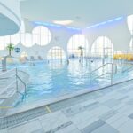 Hallenbad mit mehreren Personen, Liegestühlen, dekorativen Palmen, großen runden Fenstern und einer Wasserrutsche in einem hellen Raum.