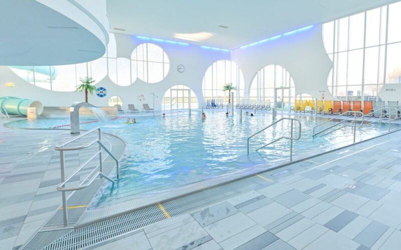 Hallenbad mit mehreren Personen, Liegestühlen, dekorativen Palmen, großen runden Fenstern und einer Wasserrutsche in einem hellen Raum.