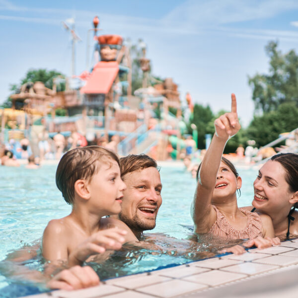 Zwei Erwachsene und zwei Kinder befinden sich in einem Schwimmbecken in der Nähe des Randes, wobei im Hintergrund ein Wasserpark und andere Menschen zu sehen sind. Ein Kind zeigt auf etwas außerhalb des Rahmens.