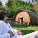 Ein Mann in einem weißen Gewand entspannt sich im Freien und blickt auf eine Frau, die am Eingang eines großen, tonnenförmigen Holzgebäudes inmitten von Grün steht.