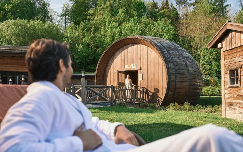 Ein Mann in einem weißen Gewand entspannt sich im Freien und blickt auf eine Frau, die am Eingang eines großen, tonnenförmigen Holzgebäudes inmitten von Grün steht.