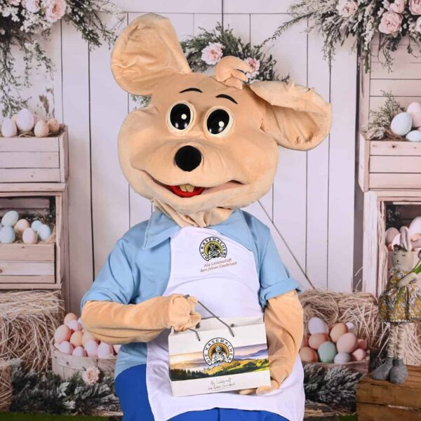 Eine Person in einem großen Mäusekostüm mit blauem Hemd und Schürze, die eine Schachtel in der Hand hält, sitzt vor einer pastellfarbenen Osterdekoration mit Eiern und Blumen.