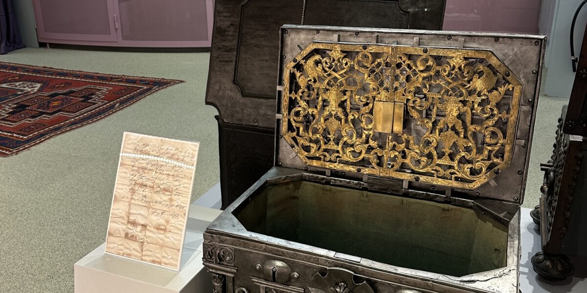 Eine verzierte Metalltruhe mit geöffnetem Deckel ist in einer Museumsausstellung zu sehen, mit historischen Schlössern an der Wand und einer Informationstafel neben der Truhe.