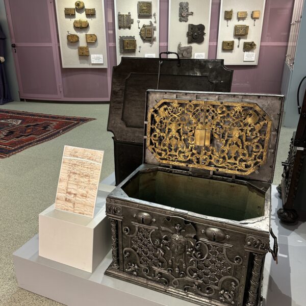 Eine verzierte Metalltruhe mit geöffnetem Deckel ist in einer Museumsausstellung zu sehen, mit historischen Schlössern an der Wand und einer Informationstafel neben der Truhe.