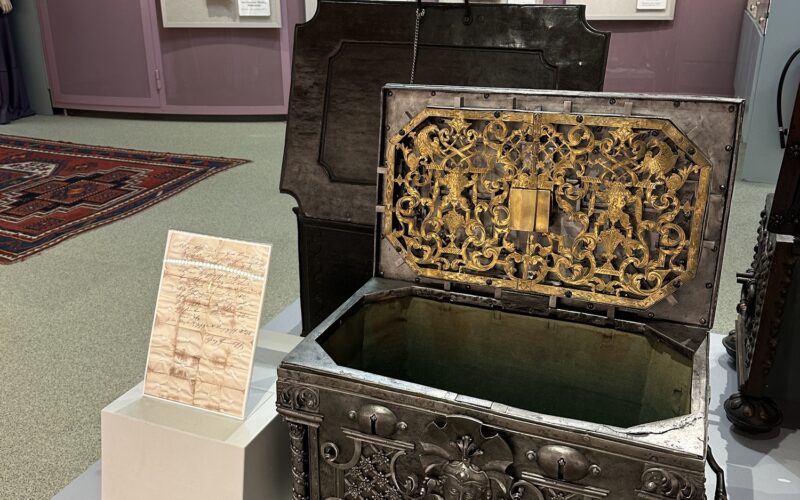 Eine verzierte Metalltruhe mit geöffnetem Deckel ist in einer Museumsausstellung zu sehen, mit historischen Schlössern an der Wand und einer Informationstafel neben der Truhe.