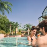 Zwei Menschen stehen an einem sonnigen Tag in einem Freibad in der Nähe eines Glasgebäudes und von Palmen, lächeln und halten Getränke in der Hand.