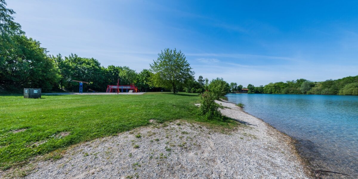 Ein grasbewachsener Park mit einem Spielplatz in der Nähe eines ruhigen Sees, gesäumt von Bäumen und einem felsigen Ufer unter einem klaren blauen Himmel.