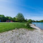 Ein grasbewachsener Park mit einem Spielplatz in der Nähe eines ruhigen Sees, gesäumt von Bäumen und einem felsigen Ufer unter einem klaren blauen Himmel.
