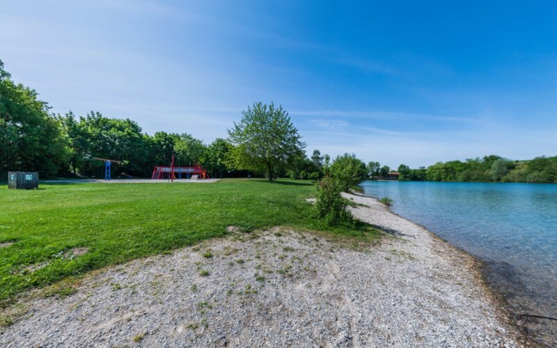 Ein grasbewachsener Park mit einem Spielplatz in der Nähe eines ruhigen Sees, gesäumt von Bäumen und einem felsigen Ufer unter einem klaren blauen Himmel.