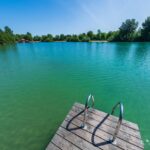 Ein hölzerner Steg mit Metallgeländern erstreckt sich über einen ruhigen, türkisfarbenen See, der von Bäumen umgeben ist, unter einem klaren blauen Himmel.