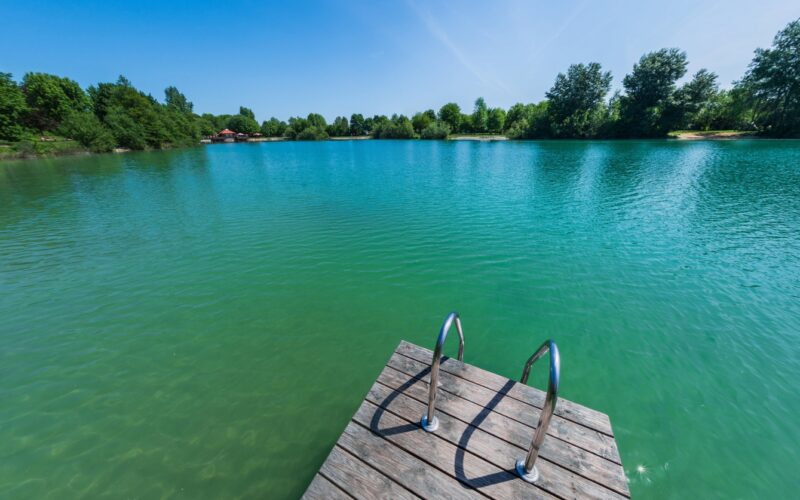 Ein hölzerner Steg mit Metallgeländern erstreckt sich über einen ruhigen, türkisfarbenen See, der von Bäumen umgeben ist, unter einem klaren blauen Himmel.