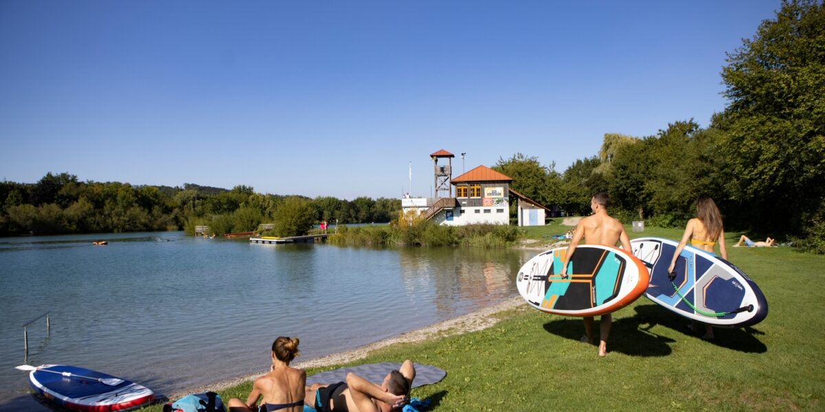 Zwei Personen tragen Paddelbretter in Richtung eines ruhigen Sees, während sich zwei andere am grasbewachsenen Ufer entspannen. Im Hintergrund sind ein Gebäude und Bäume unter einem klaren blauen Himmel zu sehen.