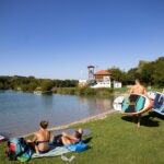 Zwei Personen tragen Paddelbretter in Richtung eines ruhigen Sees, während sich zwei andere am grasbewachsenen Ufer entspannen. Im Hintergrund sind ein Gebäude und Bäume unter einem klaren blauen Himmel zu sehen.