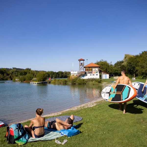 Zwei Personen tragen Paddelbretter in Richtung eines ruhigen Sees, während sich zwei andere am grasbewachsenen Ufer entspannen. Im Hintergrund sind ein Gebäude und Bäume unter einem klaren blauen Himmel zu sehen.