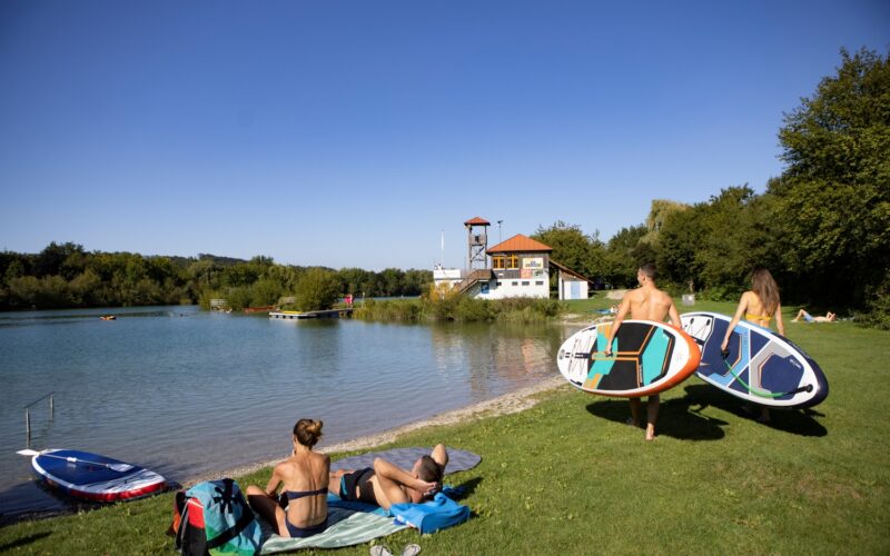 Zwei Personen tragen Paddelbretter in Richtung eines ruhigen Sees, während sich zwei andere am grasbewachsenen Ufer entspannen. Im Hintergrund sind ein Gebäude und Bäume unter einem klaren blauen Himmel zu sehen.
