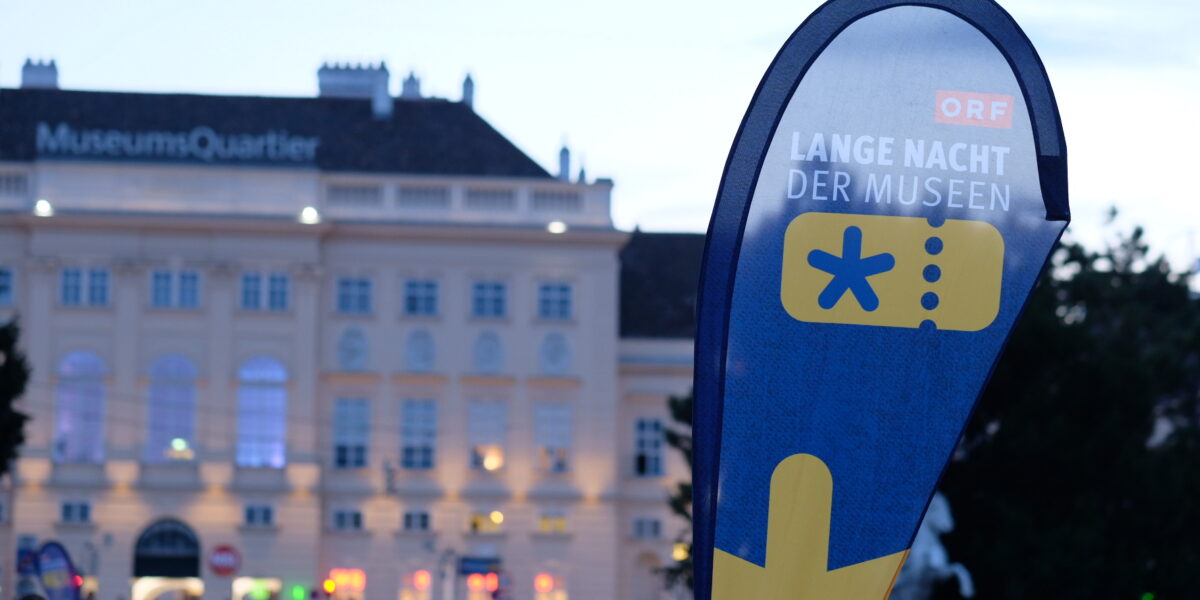 Vor dem MuseumQuartier steht ein Schild mit der Aufschrift "Lange Nacht der Museen", auf dem sich am Abend Menschen versammeln.