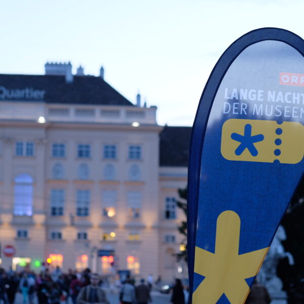 Vor dem MuseumQuartier steht ein Schild mit der Aufschrift "Lange Nacht der Museen", auf dem sich am Abend Menschen versammeln.