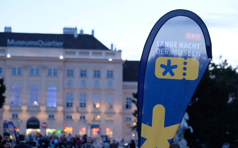 Vor dem MuseumQuartier steht ein Schild mit der Aufschrift "Lange Nacht der Museen", auf dem sich am Abend Menschen versammeln.