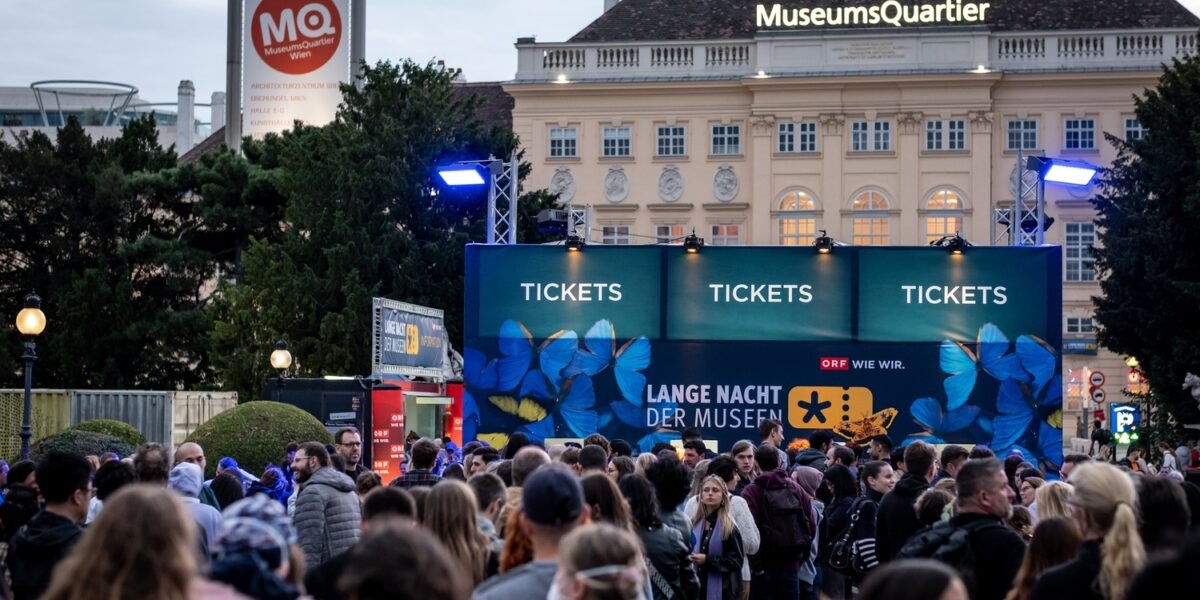 Eine große Menschenmenge versammelt sich vor den Ticketschaltern für die Lange Nacht der Museen" im MuseumsQuartier, Wien, im Hintergrund sind die Veranstaltungsschilder und Gebäude zu sehen.