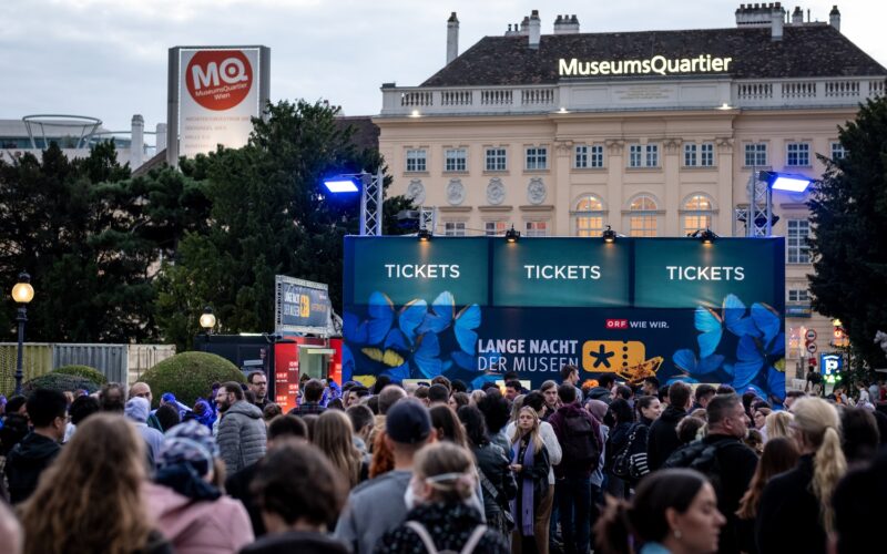 Eine große Menschenmenge versammelt sich vor den Ticketschaltern für die Lange Nacht der Museen" im MuseumsQuartier, Wien, im Hintergrund sind die Veranstaltungsschilder und Gebäude zu sehen.