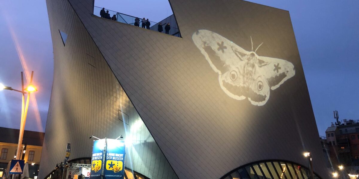 Ein modernes, kantiges Gebäude in der Abenddämmerung, auf dessen Wand ein großes Mottenbild projiziert wird; Menschen stehen auf der Dachterrasse.