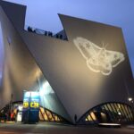 Ein modernes, kantiges Gebäude in der Abenddämmerung, auf dessen Wand ein großes Mottenbild projiziert wird; Menschen stehen auf der Dachterrasse.
