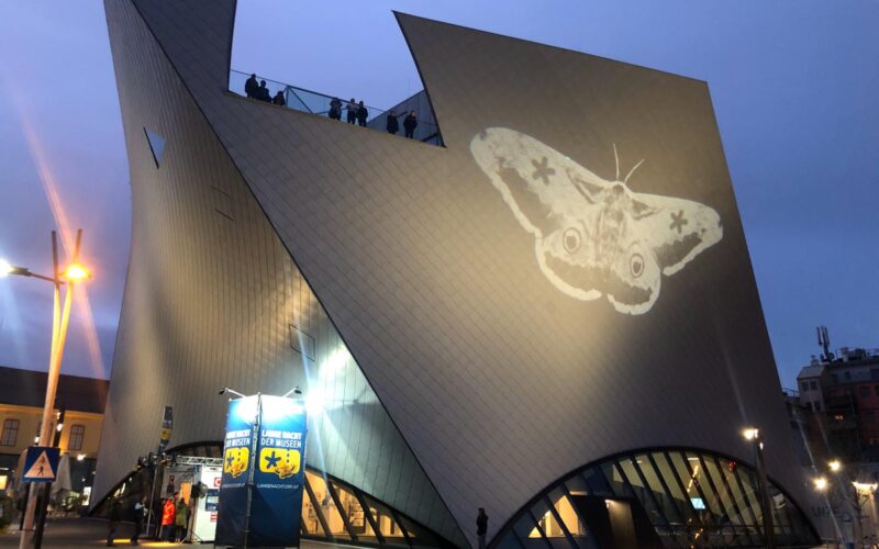 Ein modernes, kantiges Gebäude in der Abenddämmerung, auf dessen Wand ein großes Mottenbild projiziert wird; Menschen stehen auf der Dachterrasse.
