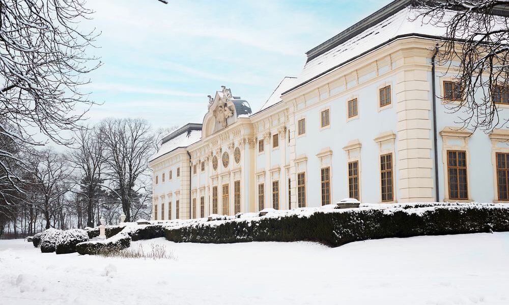 Ein großes, verziertes weißes Gebäude mit hohen Fenstern und dekorativen Details ist umgeben von schneebedecktem Boden und Bäumen unter einem bewölkten Himmel.