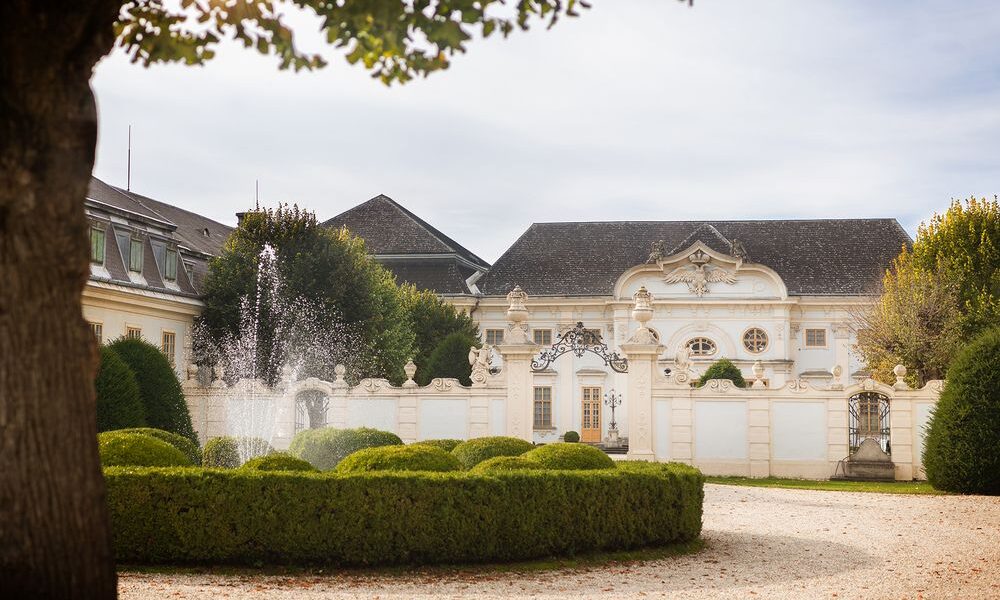 Elegantes weißes Herrenhaus mit verschnörkelter Architektur, einem dekorativen schmiedeeisernen Tor, gepflegten Hecken und einem Wasserbrunnen im Vordergrund, unter einem teilweise bewölkten Himmel.