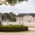 Elegantes weißes Herrenhaus mit verschnörkelter Architektur, einem dekorativen schmiedeeisernen Tor, gepflegten Hecken und einem Wasserbrunnen im Vordergrund, unter einem teilweise bewölkten Himmel.