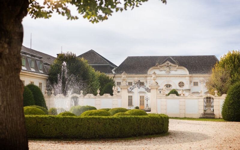 Elegantes weißes Herrenhaus mit verschnörkelter Architektur, einem dekorativen schmiedeeisernen Tor, gepflegten Hecken und einem Wasserbrunnen im Vordergrund, unter einem teilweise bewölkten Himmel.
