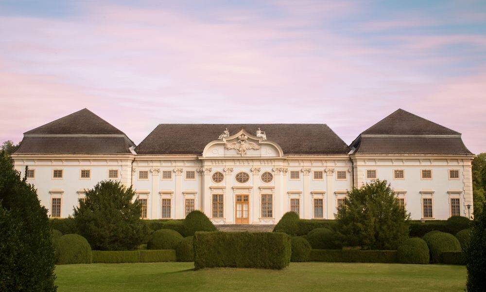 Ein großer, symmetrischer weißer Palast mit kunstvollen Details und gepflegten Gärten vor einem rosa-blauen Himmel.