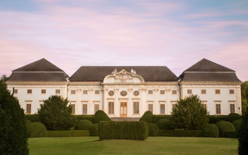 Ein großer, symmetrischer weißer Palast mit kunstvollen Details und gepflegten Gärten vor einem rosa-blauen Himmel.