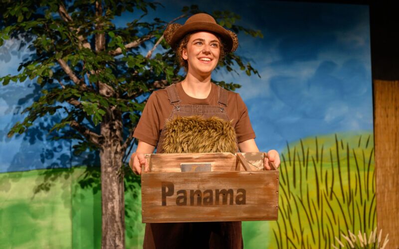 Eine Person in brauner Latzhose und Hut steht im Freien und hält eine Holzkiste mit der Aufschrift "Panama" in der Hand. Im Hintergrund sind ein Baum und eine gemalte Landschaft zu sehen.