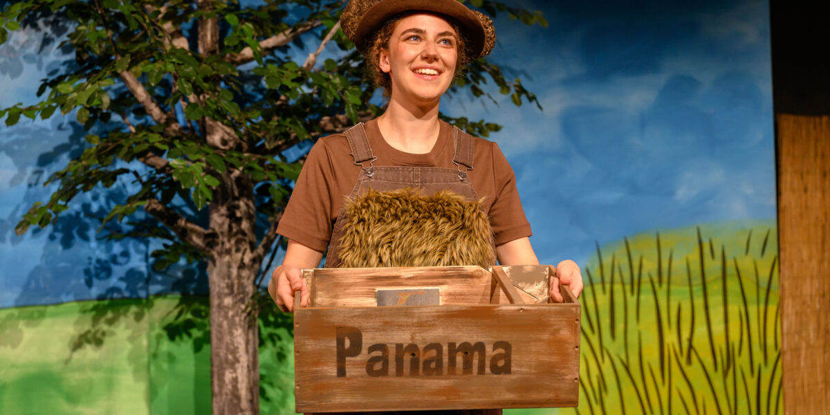 Eine Person mit braunem Hut und Latzhose hält eine Holzkiste mit der Aufschrift "Panama" auf der Bühne, vor der ein gemalter Baum und eine Landschaft zu sehen sind.