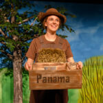 Eine Person mit braunem Hut und Latzhose hält eine Holzkiste mit der Aufschrift "Panama" auf der Bühne, vor der ein gemalter Baum und eine Landschaft zu sehen sind.