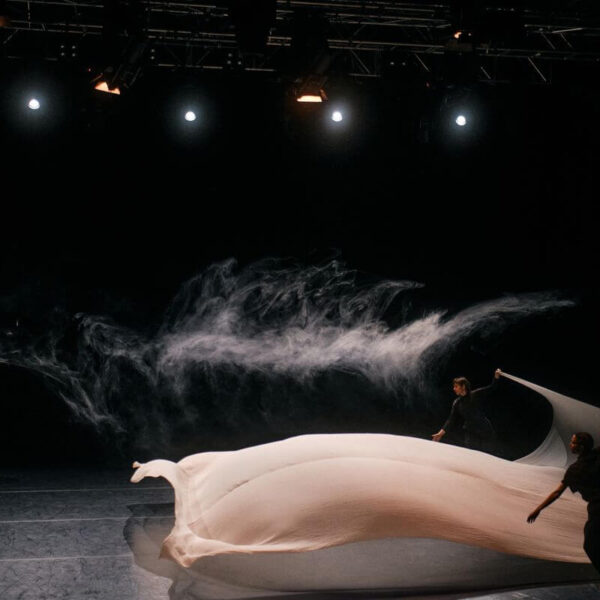 Zwei Personen manipulieren einen großen, fließenden weißen Stoff auf einer dunklen Bühne, während im Hintergrund Rauch aufgewirbelt wird, der von der Bühnenbeleuchtung beleuchtet wird.
