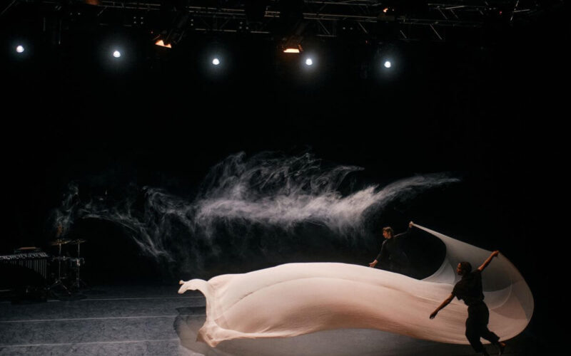 Zwei Personen manipulieren einen großen, fließenden weißen Stoff auf einer dunklen Bühne, während im Hintergrund Rauch aufgewirbelt wird, der von der Bühnenbeleuchtung beleuchtet wird.