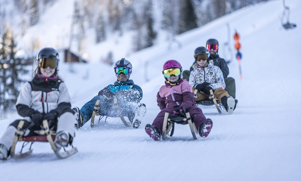Fünf Personen in Winterausrüstung und mit Helmen schlittern einen verschneiten Hügel hinunter, umgeben von Bäumen und entfernten Skiliftstangen im Hintergrund.