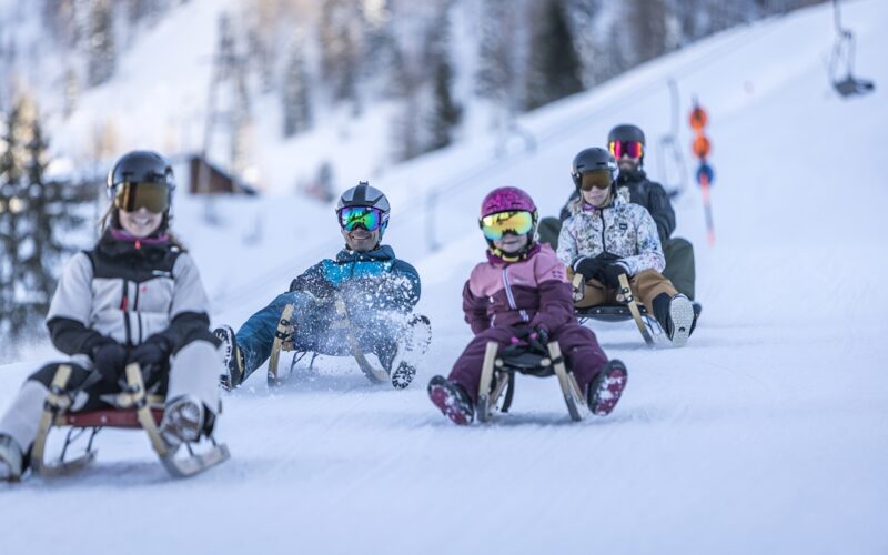Fünf Personen in Winterausrüstung und mit Helmen schlittern einen verschneiten Hügel hinunter, umgeben von Bäumen und entfernten Skiliftstangen im Hintergrund.