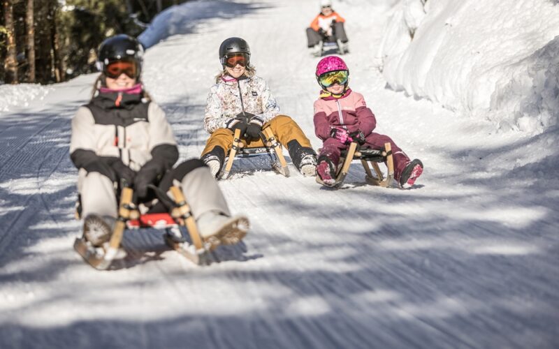 Vier Personen fahren mit Helmen und Winterkleidung auf Holzschlitten einen verschneiten, von Bäumen gesäumten Weg hinunter.
