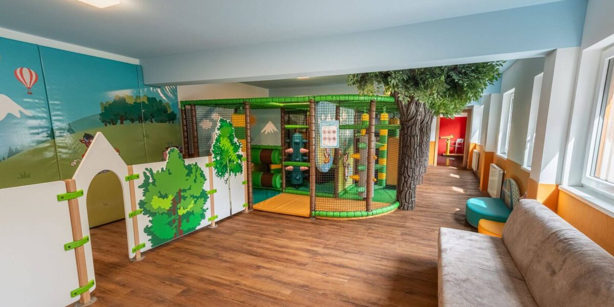 Ein Indoor-Spielbereich für Kinder mit einem kleinen Klettergerüst, weichen Barrieren, einem künstlichen Baum, einem Wandgemälde und einer Couch auf einem Holzfußboden.