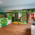 Ein Indoor-Spielbereich für Kinder mit einem kleinen Klettergerüst, weichen Barrieren, einem künstlichen Baum, einem Wandgemälde und einer Couch auf einem Holzfußboden.