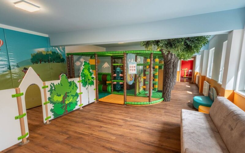 Ein Indoor-Spielbereich für Kinder mit einem kleinen Klettergerüst, weichen Barrieren, einem künstlichen Baum, einem Wandgemälde und einer Couch auf einem Holzfußboden.