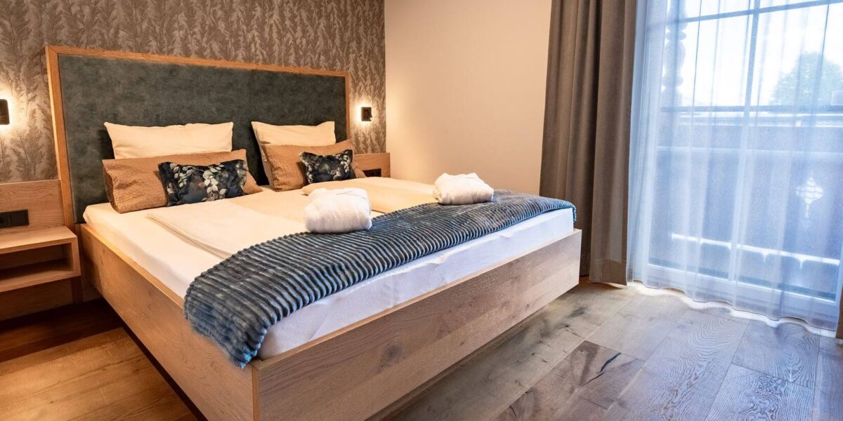 Modernes Hotelzimmer mit einem großen Holzbett, zwei Kissen, gefalteten Handtüchern und einer blauen Überwurfdecke. Das Zimmer hat Holzböden, gemusterte Tapeten und durchsichtige Vorhänge vor einem Fenster.