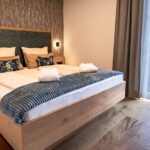 Modernes Hotelzimmer mit einem großen Holzbett, zwei Kissen, gefalteten Handtüchern und einer blauen Überwurfdecke. Das Zimmer hat Holzböden, gemusterte Tapeten und durchsichtige Vorhänge vor einem Fenster.