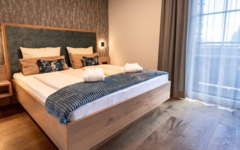 Modernes Hotelzimmer mit einem großen Holzbett, zwei Kissen, gefalteten Handtüchern und einer blauen Überwurfdecke. Das Zimmer hat Holzböden, gemusterte Tapeten und durchsichtige Vorhänge vor einem Fenster.